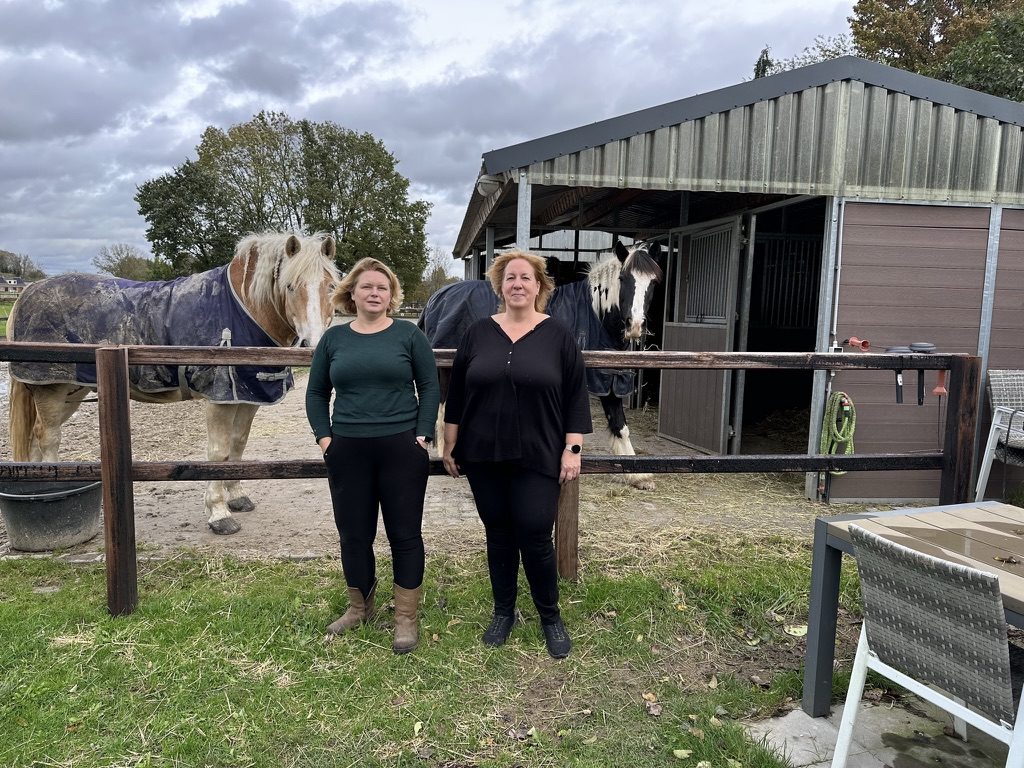 Jessica en Chantal staan voor de stal met de paarden achter het hek