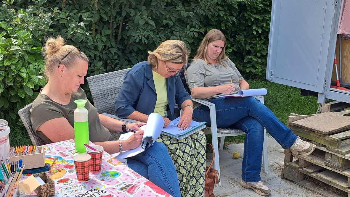 Drie cursisten zitten buiten in de tuin te schrijven in hun werkboek